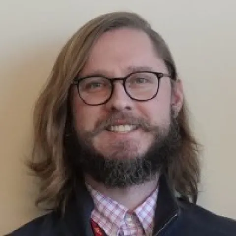 White man with shoulder length brown hair, a beard and black glasses smiling