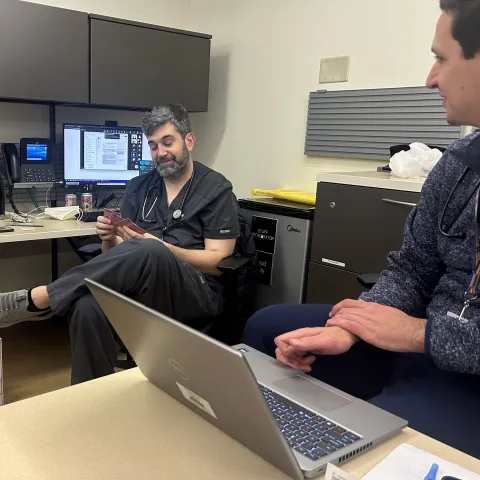 Two men sit at desks with computers, talking 