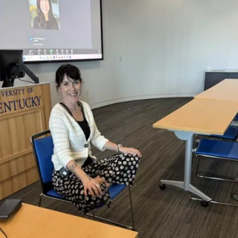 A smiling woman sits at a desk in a University of Kentucky classroom with a video call on the screen. 