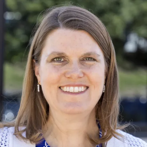 April Stauffer wears a blue patterned blouse and knitted white cardigan while smiling for a photo. 