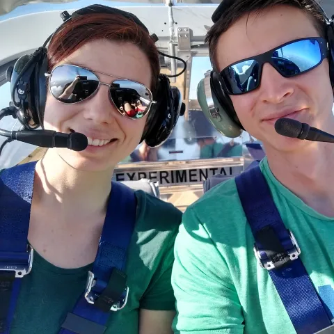 Leigh and Her Husband Wes smiling in an airplane while flying. They both have mics and shades on. 