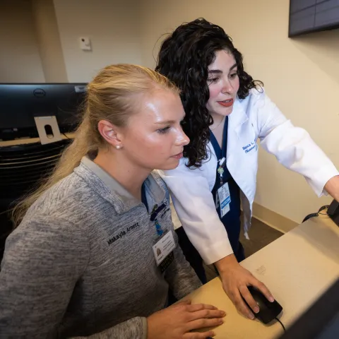 A female student being guided through a computer process by a woman in a white coat.