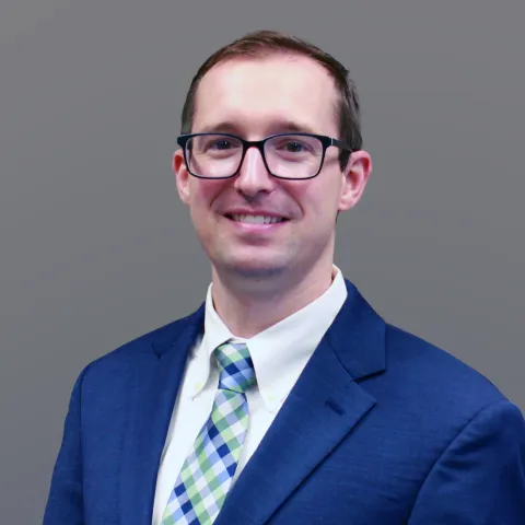Andrew Bishop, 2025 graduate of CT Surgery integrated residency, is in blue blazer, white shirt, and plaid tie. He wears hiorn-rimmed glasses