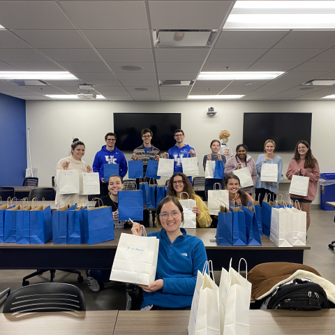 various students hold up blue and white gift bags from a care package packing event