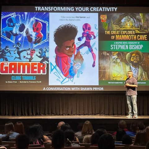 Pryor giving a presentation on a stage about graphic novels, with two book covers projected on the slides. They are titled "The Gamer, Clone Trouble," and "The Great Explorer of Mammoth Cave "