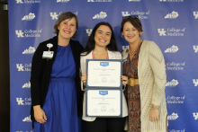 Awardee and presenters hold two certificates and pose for a photo in front of blue backgroudn
