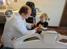 Dr. Arnold kneels beside a hospital bed, playfully interacting with a smiling toddler holding a stethoscope.