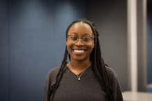 Young lady in black top standing in front of blue background, smiling