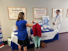Two women lean near a table of informational pamphlets. One of them speaks to the doctor who stands nearby.