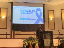 Pastor Gaines stands in front of a screen advertising colon cancer awareness