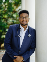 Kidus poses in a suit with a stethoscope around his neck, leaned against a pillar.