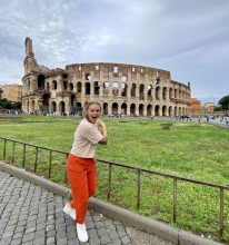 Ward poses excitedly in front of the Colosseum in Rome, with a cloudy sky and lush green grass in the background.