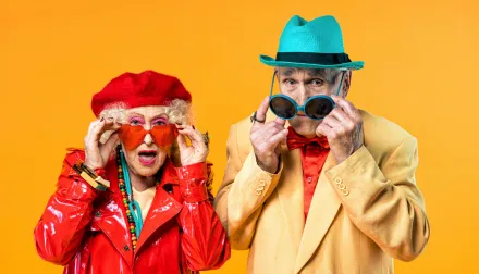Elderly couple is dressed up in colorful, fancy clothes against an orange background. 