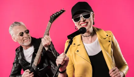 Elderly couple are playing rock music on instruments against a hot pink background.