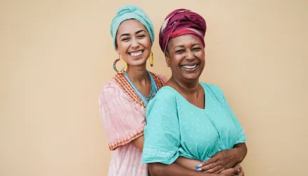 An elderly woman is smiling and hugging her adult daughter against a tan background. 