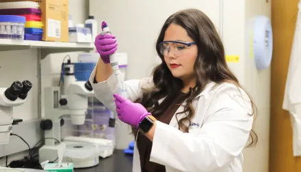 Katie Land, PhD student, wears a white coat and PPE and uses equipment in a lab setting