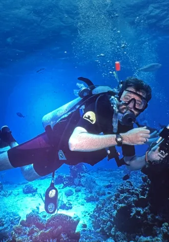 Scuba diver in clear blue ocean, holding a camera, surrounded by coral and fish. 