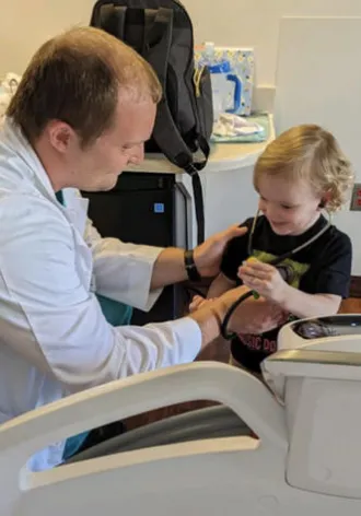 Andre Arnold, dressed in his white coat, helps a child hear their own heart beat using his stethoscope.