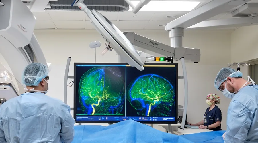 diagnostic room with three providers and patient laying on bed; lots of radiologic equipment; screen with images of patient's brain