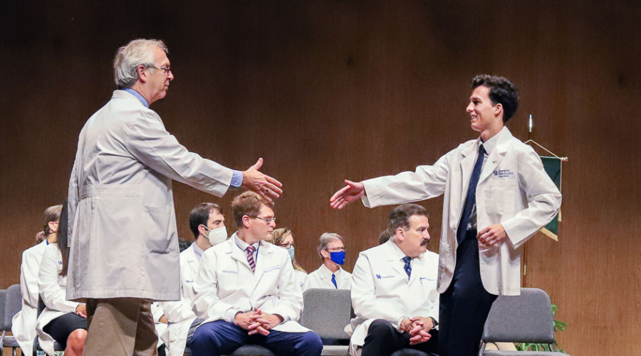 two white coats, presumably one an instructor and one a student, are extending hands to shake on a stage