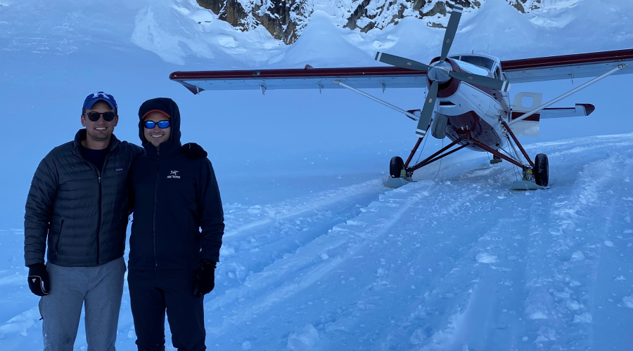 Sam Hughes standing with a friend in the snow in front of a mountain and parked airplane