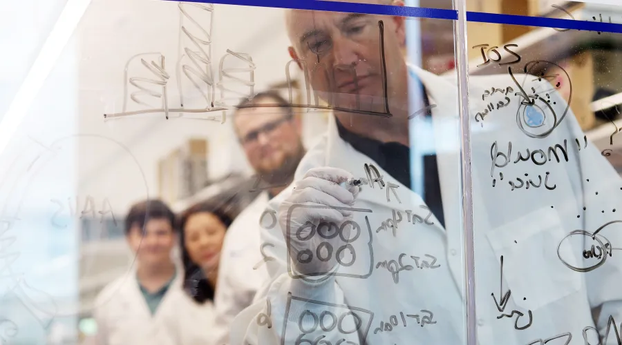 dr. terry hinds writing on a transparent dry erase board with learners watching from behind him