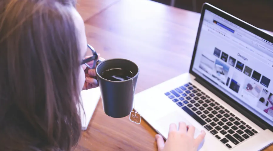 A woman holding a cup of tea, working on a laptop.