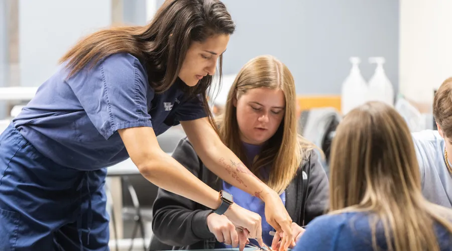 Standing woman in medical scrubs shows sitting student suturing technique in a hands-on workshop.