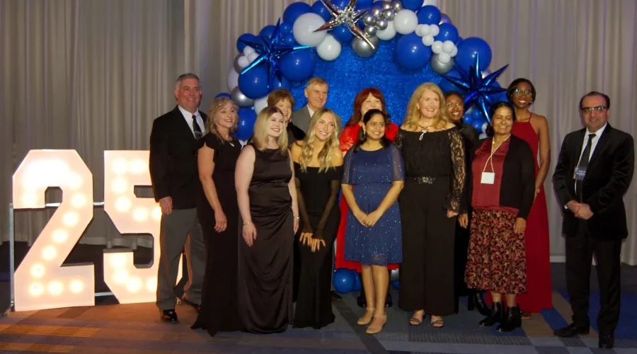 A large group of people in formal attire pose for a photo. They stand in front of a balloon arch and big, glowing numbers that say 25