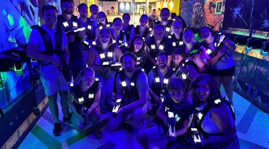 A group of people wearing glowing vests pose in an indoor laser tag arena. The room is lit with vibrant blue and yellow lights, creating an energetic and playful atmosphere.