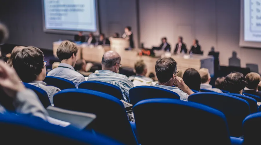A lecture hall of people. The photo is taken from the back, so you see the listeners' heads and the presenters down at the front