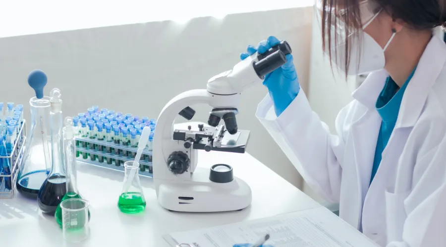 A woman in a mask looks through a microscope. Around her sits vials and tubes of liquid.