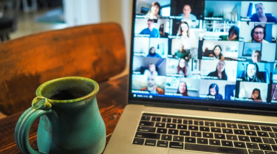 Laptop displaying a video conference with multiple participants, beside a coffee mug on a wooden table.
