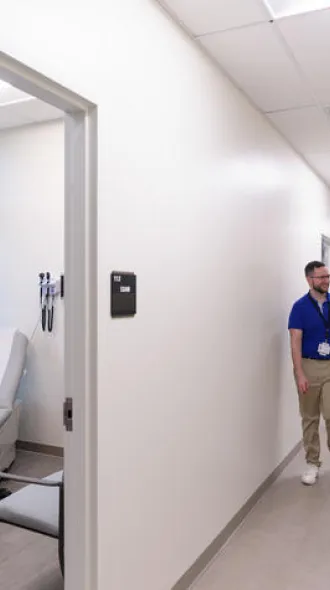 Four people speak at a hospital in two pairs. One pair speaks inside of a hospital room, looking at a computer. The other pair speaks outside in the hallway, smiling.