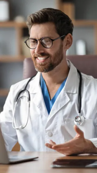 A man with dark hair, a beard, and glasses, wearing a white coat while holding a web call.