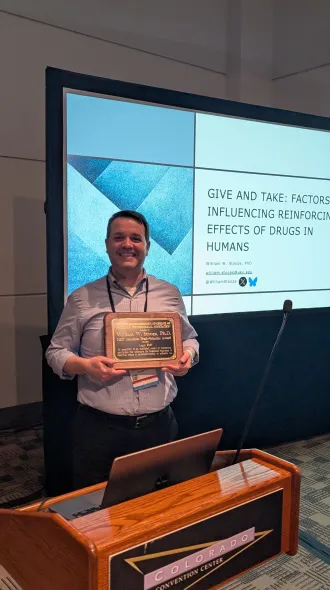 Dr. Stoops poses in front of a projected screen, holding his plaque