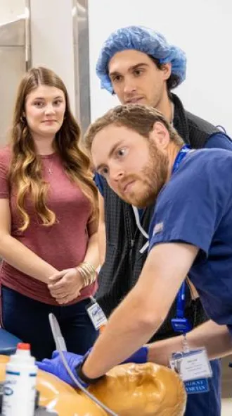 A group of five people attentively observes a medical simulation as an anesthesiology resident in scrubs demonstrates a procedure on a mannequin. The setting is clinical, with a focused and educational atmosphere.