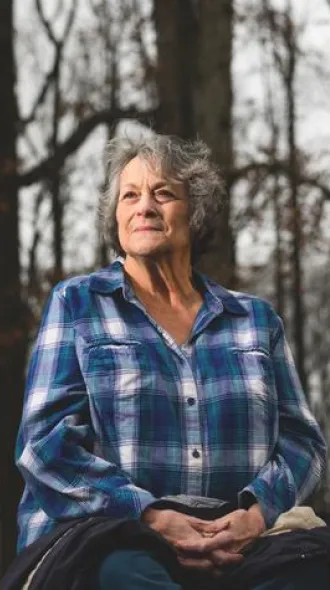 An elderly woman with short, grey hair, wearing a blue plaid shirt. She is sitting outdoors in front of a tree-line, with hands in her lap. She smiles softly into the light of the setting sun.