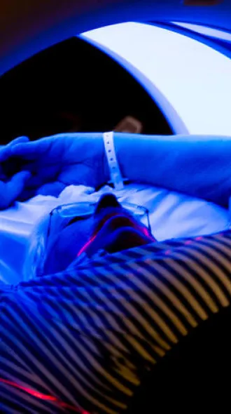 A man in a striped shirt, laying back and holding his arms over his head while receiving a CT scan.