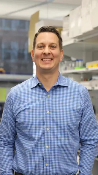 Lance Johnson in a blue checkered shirt stands in a bright laboratory. Shelves with colorful supplies and a microscope are visible, conveying a professional yet welcoming atmosphere.