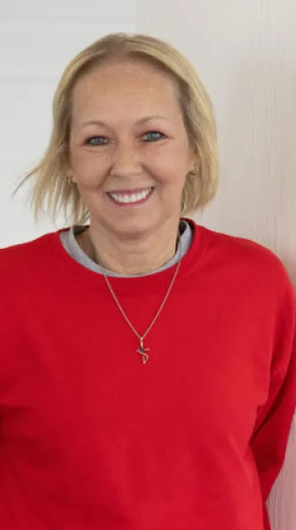 Vikki Lane, a blond woman wearing a red sweatshirt, leans against a wall and smiles. The image evokes positivity and happiness.