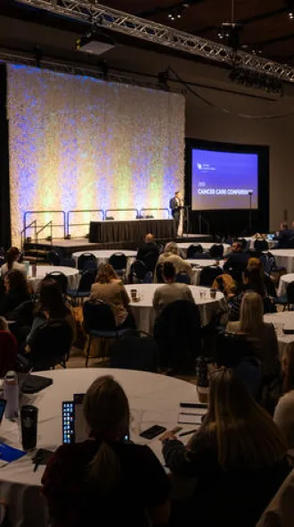 Large conference room with attendees seated at round tables facing a stage. Screens display "2023 Cancer Care Conference." The atmosphere is focused and professional.