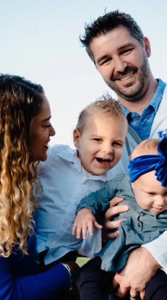 Two adults holding two young children outdoors near a wooden fence with green trees and a clear sky in the background.
