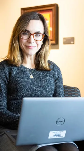 Sara Police sits in an office chair, smiling, and holding a laptop. The background features a colorful painting and an anatomical poster, creating a scholarly atmosphere.