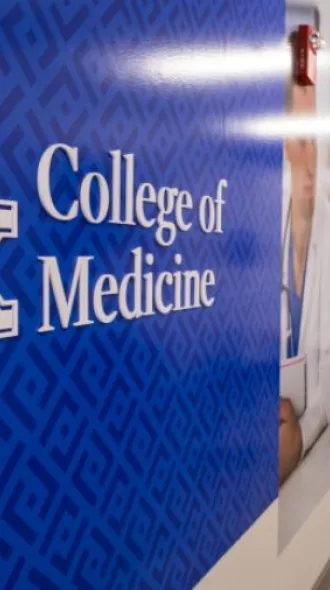 Three medical students wearing white coats walk down long hallway, UK College of Medicine logo to the left