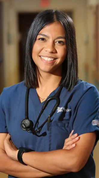 Smiling health care professional in navy scrubs with a stethoscope, standing confidently in a clinic hallway with arms crossed.