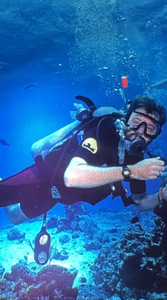Scuba diver in clear blue ocean, holding a camera, surrounded by coral and fish. 