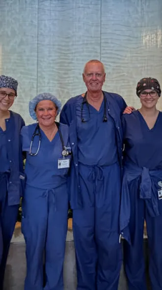 A smiling group of physicians stand in blue scrubs in front of a water wall in the UK hospital. The photo conveys positivity and community.