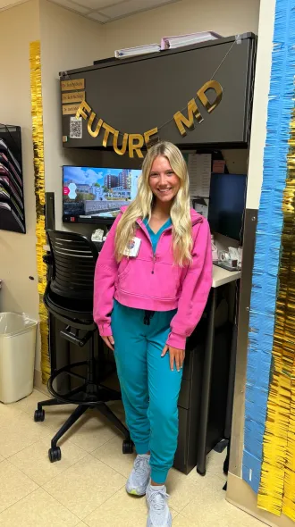 A woman in a pink hoodie and teal scrub pants smiles in an office decorated with "Future MD" banner. The room has gold and blue streamers, creating a celebratory tone.