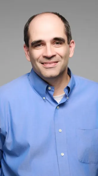 Headshot of Dr. Penaskovic, wearing a light blue button-up shirt in front of a gray background, smiling gently. 